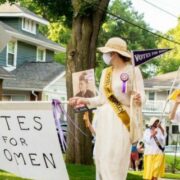 Image shows people walking down a neighborhood street in the parade. Two women in white dresses and hats and wearing masks for covid-19 hold a banner that says "Votes for Women." Participants also hold pennants and wear sashes that say "Votes for Women."