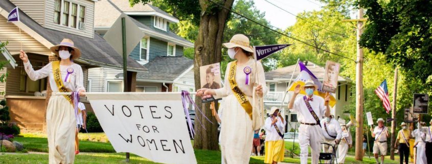 Image shows people walking down a neighborhood street in the parade. Two women in white dresses and hats and wearing masks for covid-19 hold a banner that says "Votes for Women." Participants also hold pennants and wear sashes that say "Votes for Women."